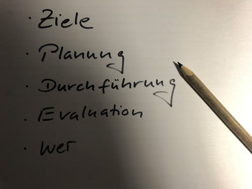 Blatt Papier mit handschriftlichen Stichpunkten: „Ziele“, „Planung“, „Durchführung“, „Evaluation“, „wer“. Rechts daneben ein Bleistift.