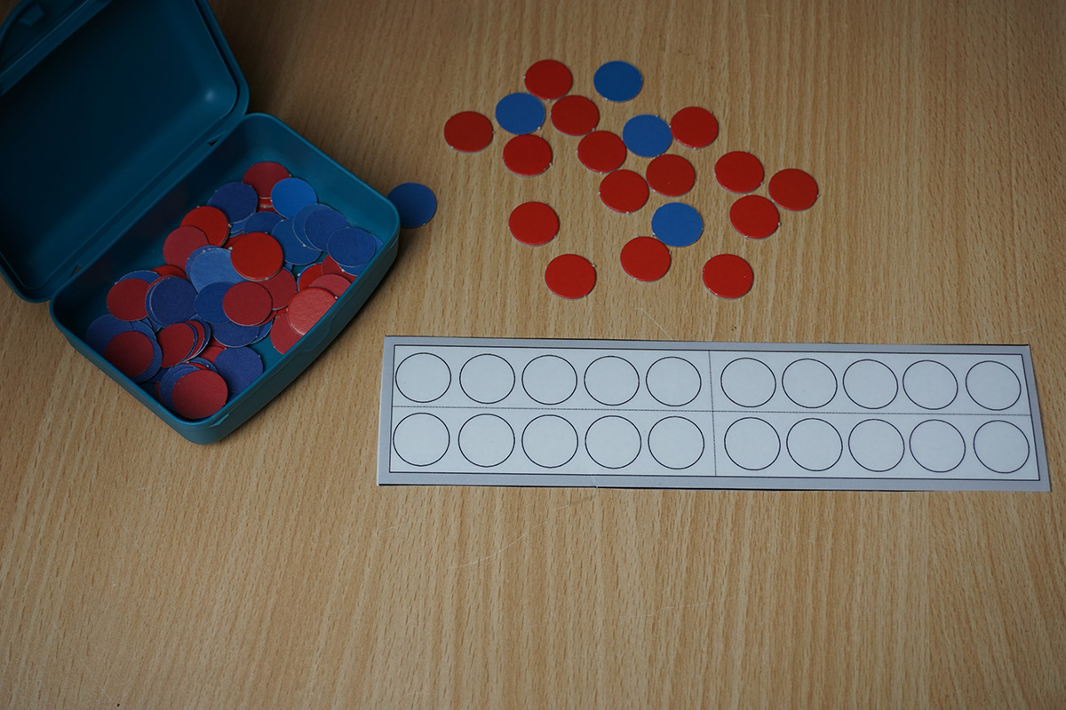Links eine Box mit roten und blauen Plättchen. Rechts daneben liegen unsortierte Plättchen. Darunter ein leeres Zwanzigerfeld (2 Reihen mit 10 leeren Kreisen, Strich nach 5 Punkten).