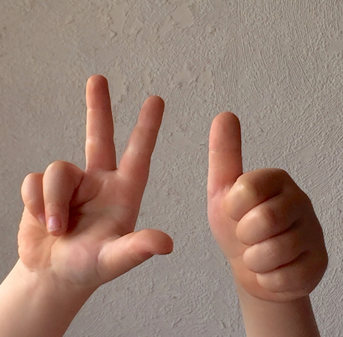 Foto von zwei Kinderhänden. Handflächen zeigen zum Betrachter. Linke Hand: Ring- und kleiner Finger zeigen in die Handinnenfläche. Die anderen Finger sind ausgestreckt. Rechte Hand: Der Daumen ist ausgestreckt. Alle anderen Finger zeigen in die Handinnenfläche.