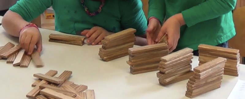 Foto: Auf einem Tisch liegen fünf Türme aus jeweils 10 Kapla-Steinen. Zwei Kinder bauen mit ihren Händen an weiteren unvollständigen Türmen. Weitere Steine liegen unsortiert in der linken Ecke.