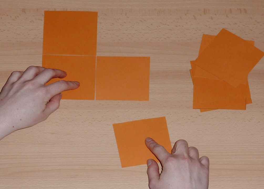 Zwei Hände verschieben große Pappquadrate und formen einen Drilling in Form des Buchstaben L. Rechts daneben liegt ein Stapel von Pappquadraten.