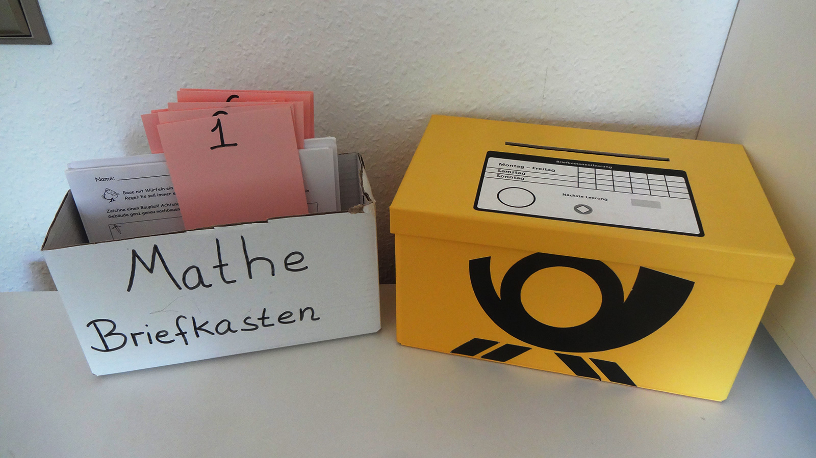Links Pappkarton mit der Aufschrift „Mathe Briefkasten“. Im Karton befinden sich rosa Zettel mit Nummerierungen, dahinter jeweils Aufgabenblätter. Rechts daneben ein gelber Pappkarton mit Deckel und dem Logo der Deutschen Post. Im Deckel befindet sich ein Schlitz.