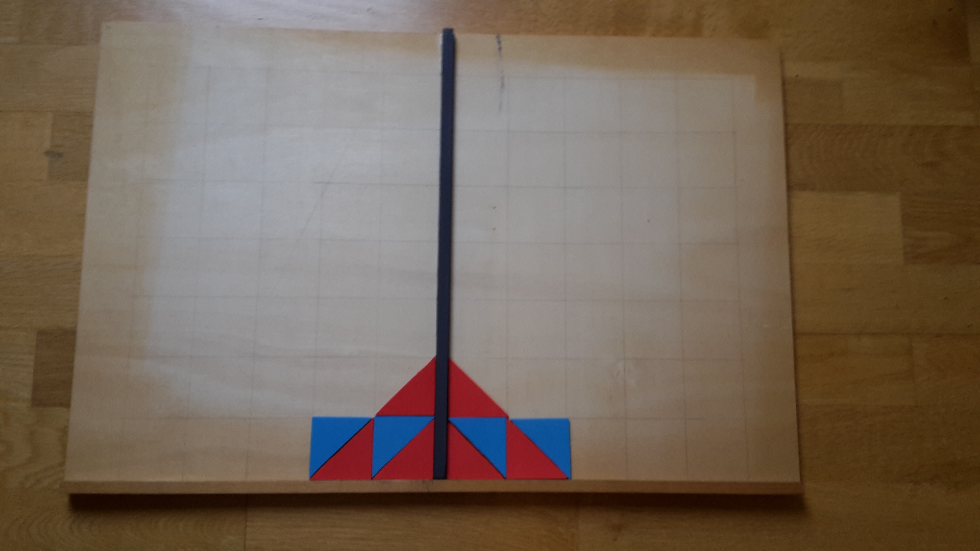 Unterlage mit Kästchenstruktur mit Spiegelachse in der Mitte. Links neben der Spiegelachse liegt ein Muster aus roten und blauen Dreiecken. Rechts neben der Spiegelachse ist es achsensymmetrisch abgebildet. Das rote Dreieck aus Abbildung 4 ist in dem Muster enthalten.